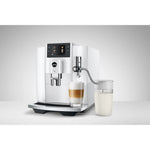 JURA Milk container EA (24313) shown connected to a white E8 coffee machine with tubing and a glass of milk foam beside it
