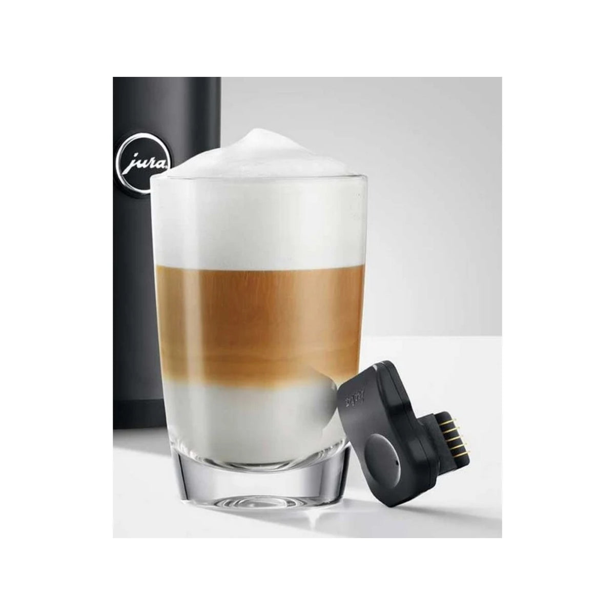JURA Bezdrôtový vysielač pre Cool Control 1 liter (24031) vedľa sklenice s vrstveným latté a časti čierneho prístroja s logom