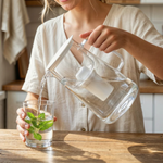 Woman pouring water from a transparent glass filter pitcher with white filter into a glass with mint on a wooden table