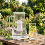 Glass filter pitcher with a white insert on a wooden table next to a glass of water with lemon and mint in an outdoor setting