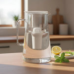Transparent glass filter pitcher with white filter cartridge on a kitchen counter next to lemon slices and mint leaves